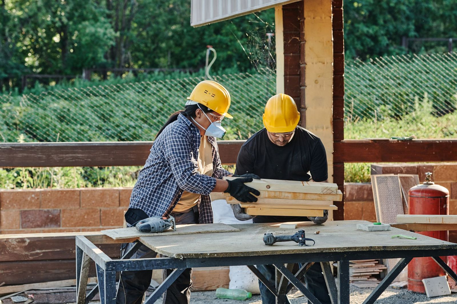 Two tradespeople in hard hats measuring and marking lumber on a site workbench