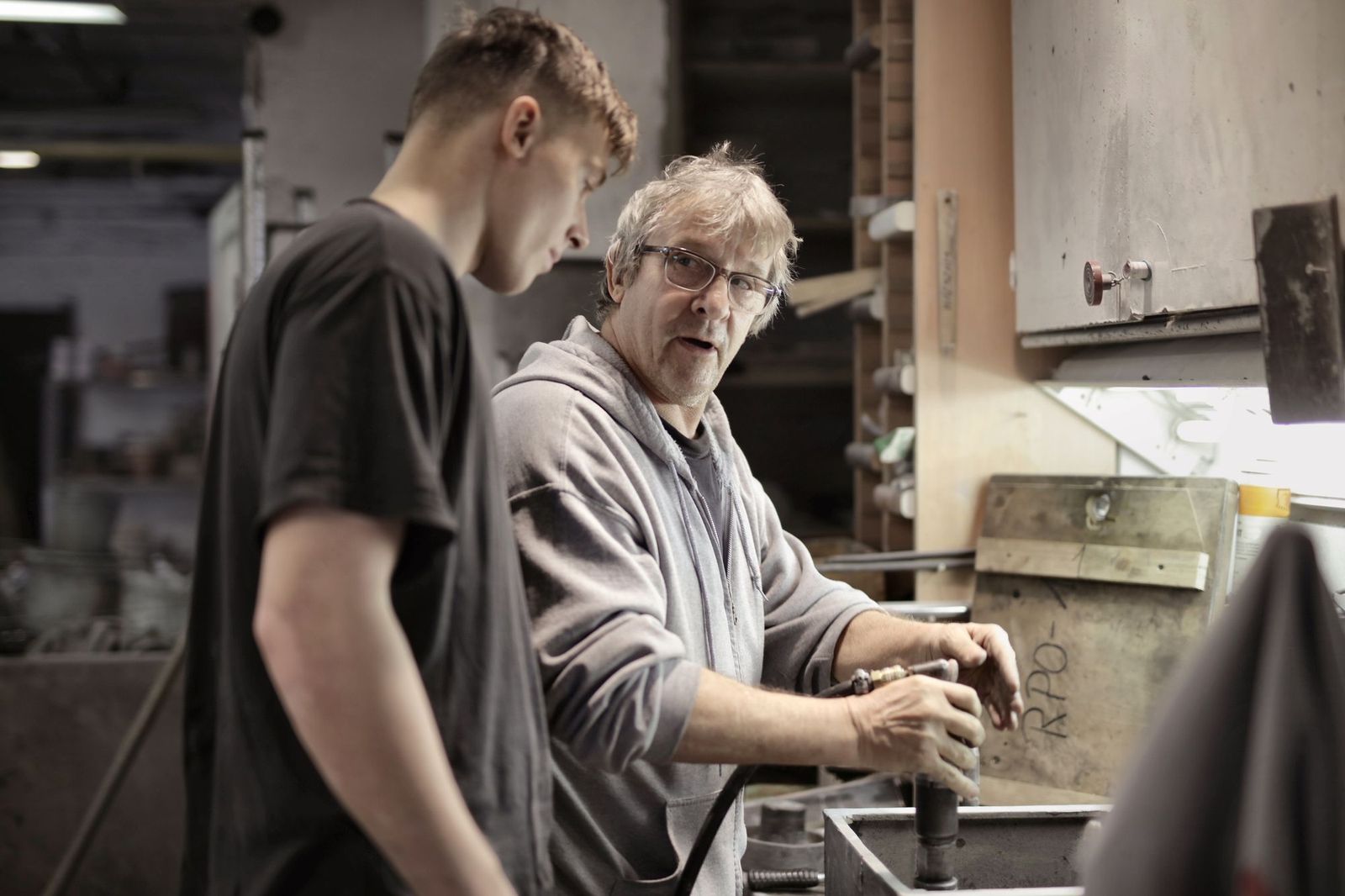 A senior tradesman showing a younger trainee how to handle a part at a workshop bench