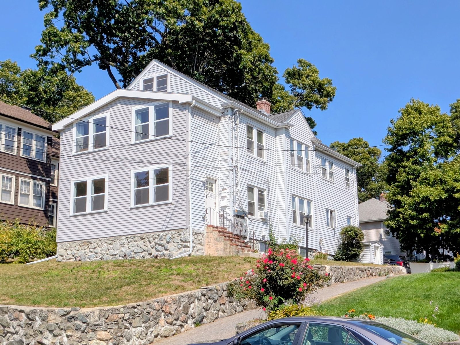 The finished Waltham house with its new two-story front addition in white vinyl siding, new windows and the stone foundation, on a sunny day