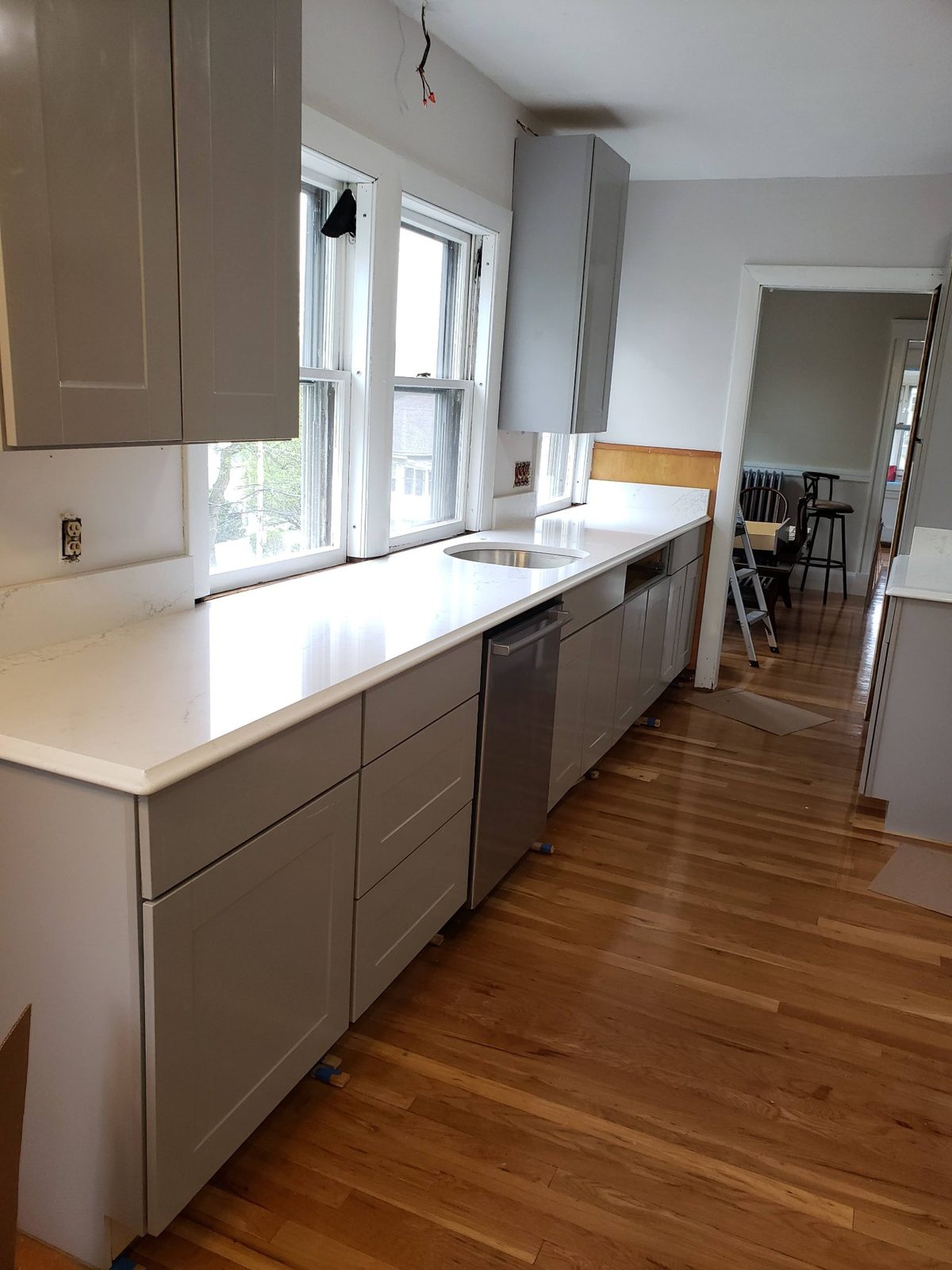 Renovated kitchen with new grey shaker cabinets, white quartz countertop, undermount sink and stainless dishwasher over refinished hardwood floors