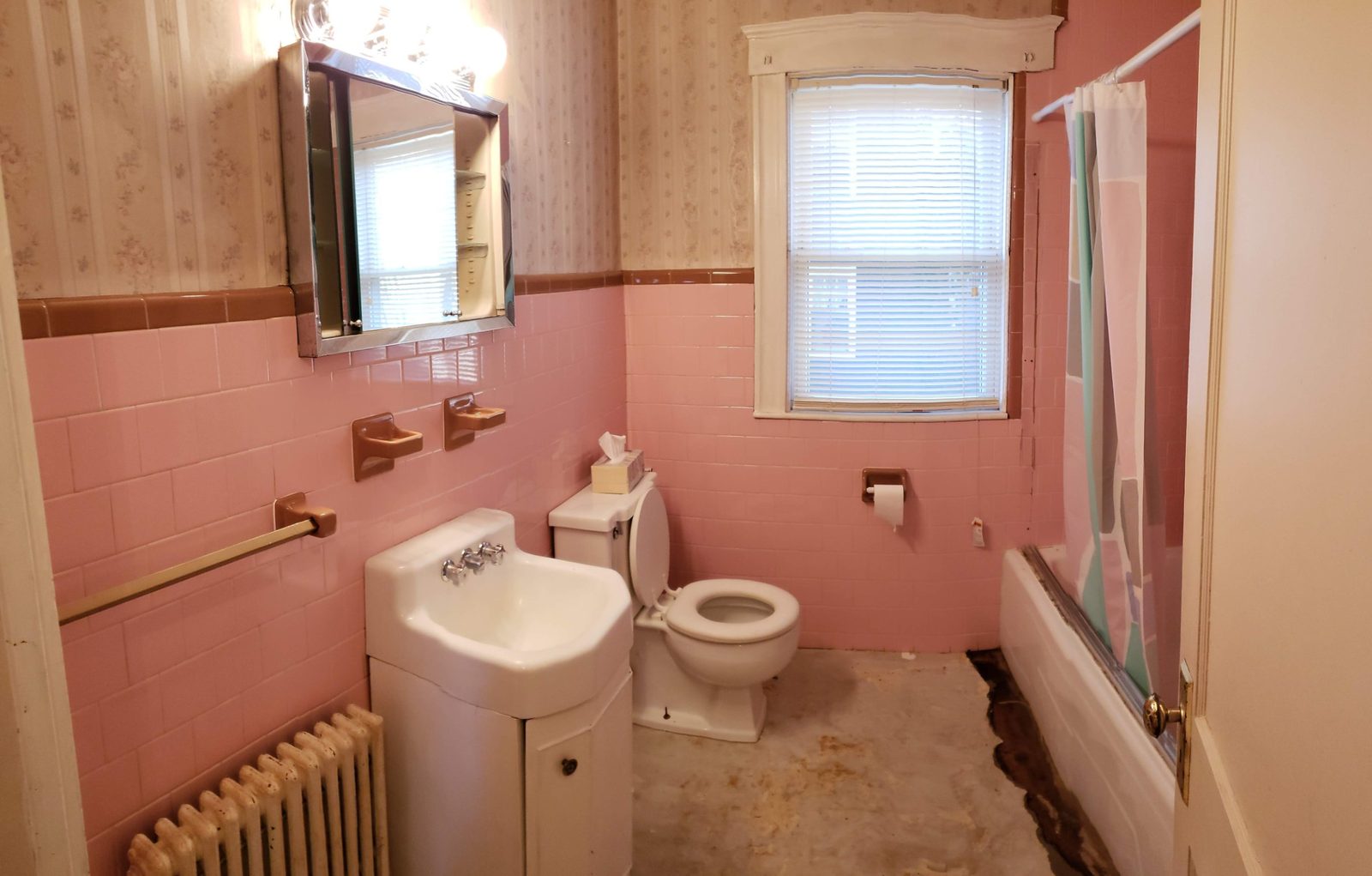 The original bathroom with pink wall tile, brown trim, wallpaper above and the floor partly torn up