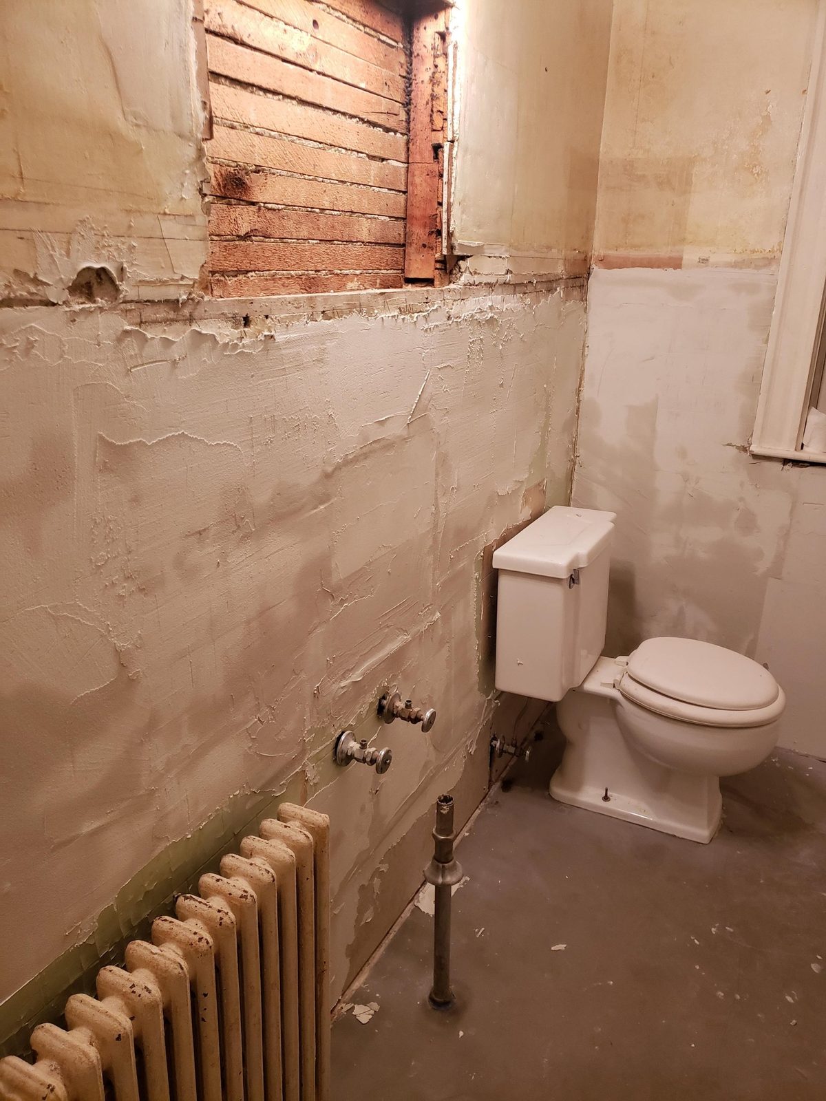 The bathroom during demolition, with tile removed, plaster patched, a wall cavity open and supply lines exposed