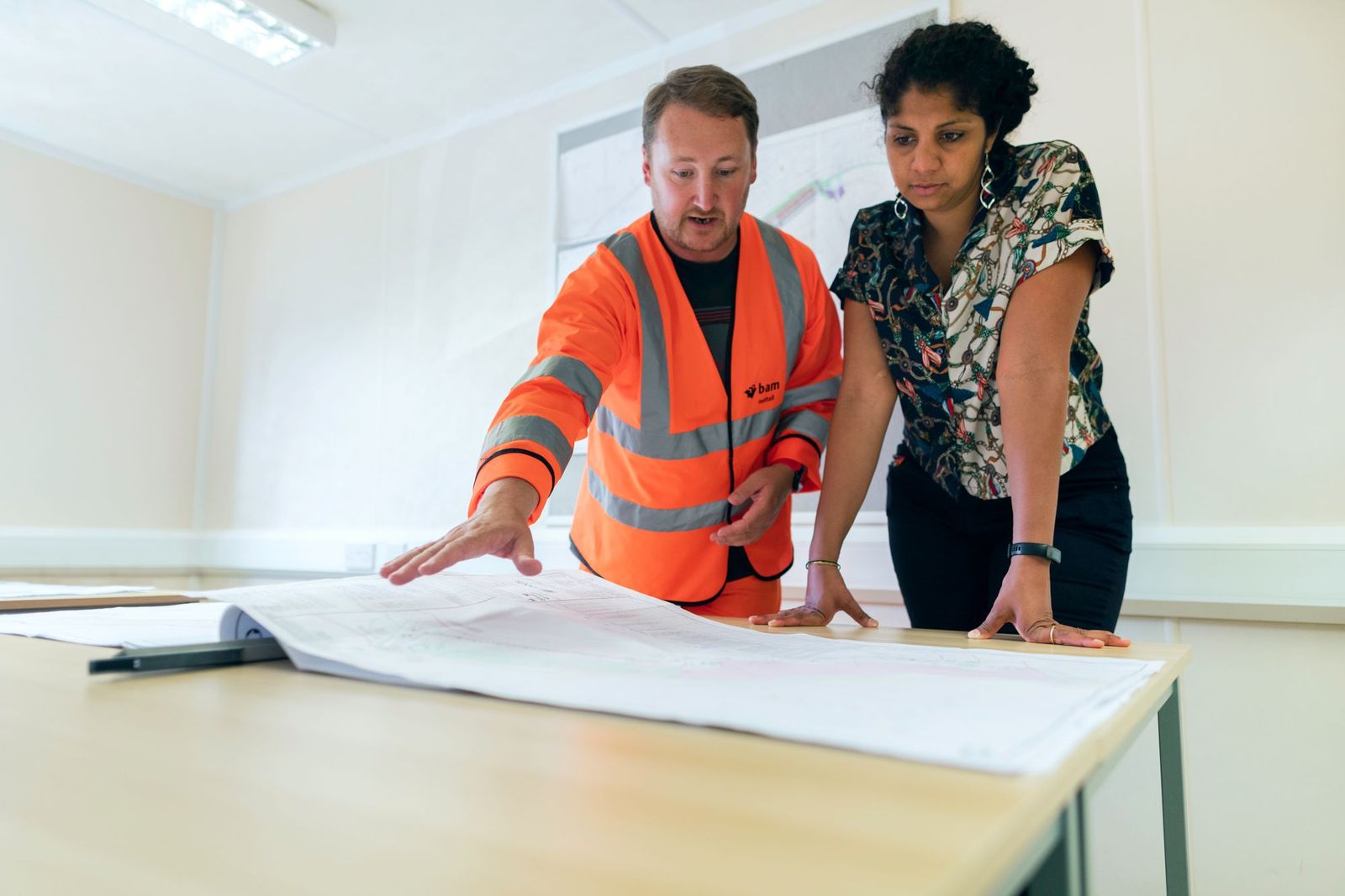 Engineer in a high-visibility jacket and a colleague reviewing construction drawings together at a table